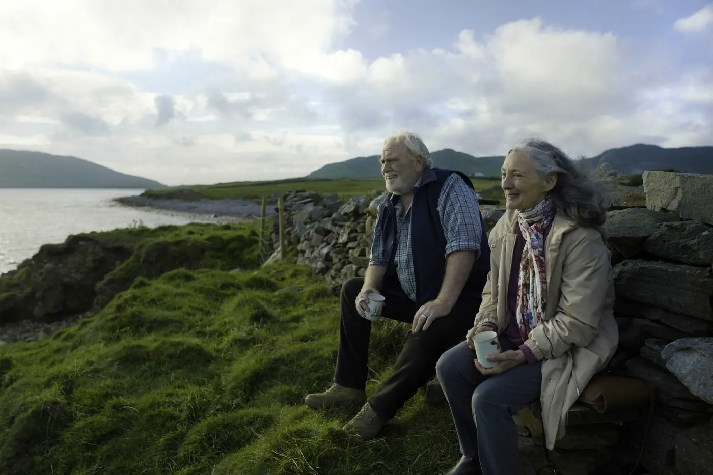 felsige Küstenlandschaft, älterer Mann und ältere Frau sitzen auf einer Bank und schauen zum Wasser