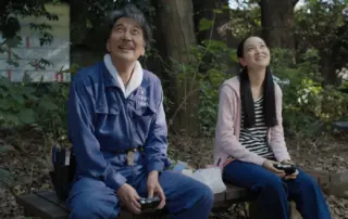 ein älterer Mann und eine junge Frau sitzen auf einer Bank unter einem großen Baum, sie lächeln, ihr Blick ist nach oben gerichtet