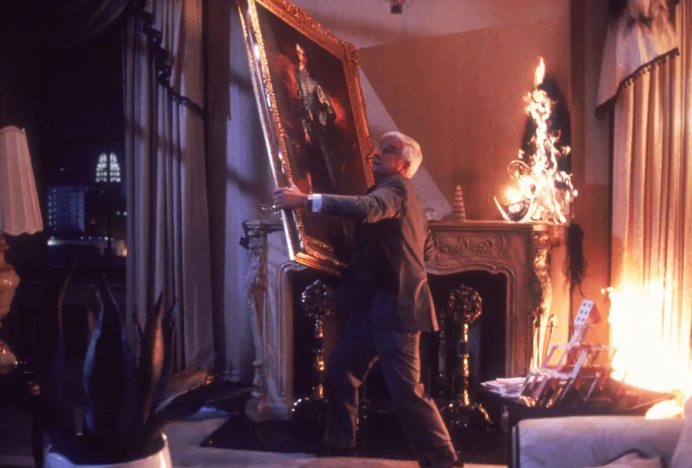 ein Kaminzimmer bei Nacht, im rechten Teil des Bildes hat ein Wandschmuck Feuer gefangen, in der Mitte des Bildes rettet Frank Drebin ein meterhohes Gemälde vor dem Feuer