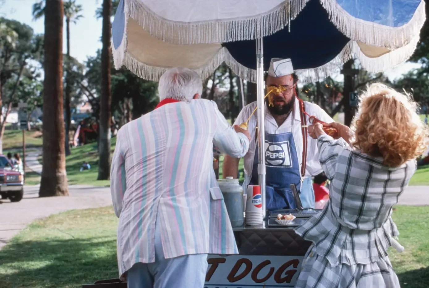 ein Hot-Dog-Verkäufer steht am Straßenrand eines begrünten Verkehrsbereiches, ein Mann und eine Frau stehen vor seinem Ständchen mit einem blauweißen Sonnenschirm und spritzen dem Verkäufer mit einer Ketschup- und einer Senfflasche ins Gesicht