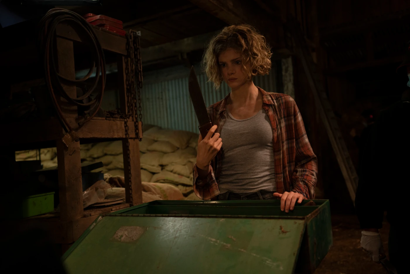 eine Frau steht in einem Lagerraum vor einer geöffneten Truhe, sie hält ein Messer in der Hand. Im Hintergrund stapeln sich braune Säcke, während das Licht gedimmt ist