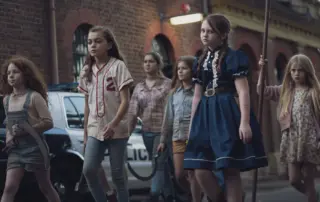 eine Gruppe von Mädchen geht eine Straße entlang, sie sind mit Hammer und Sensen bewaffnet, ihre Blicke sind grimmig. Im Hintergrund parkt ein Polizeiauto.