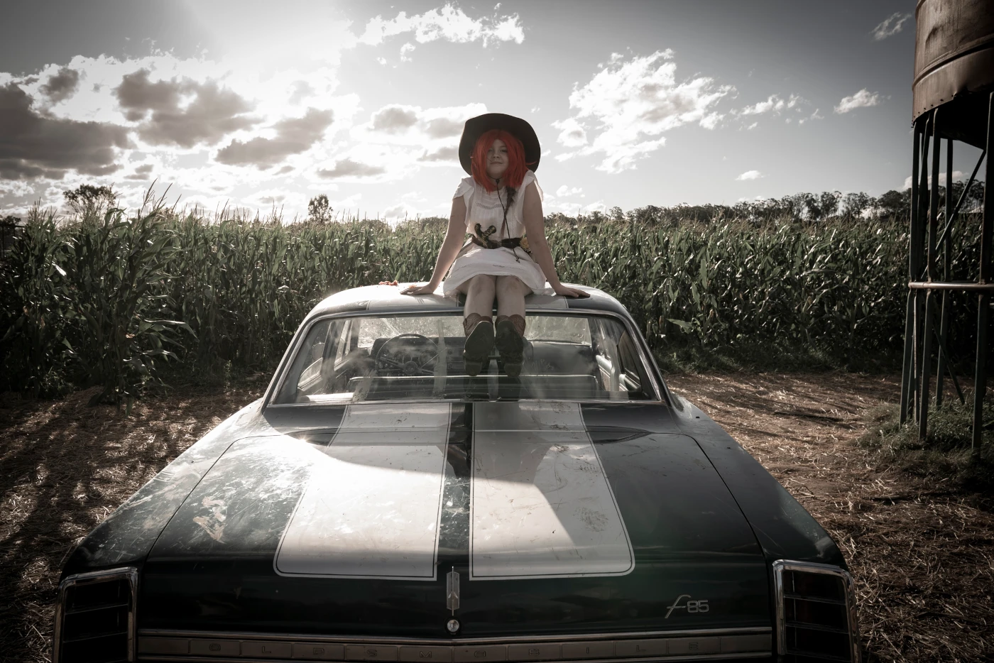 ein Mädchen mit roter Perücke, schwarzen Hut und weißem Kleid sitzt auf dem Dach eines stehenden schwarzweißen Autos, das Mädchen ist bewaffnet, im Hintergrund ist ein sonnenbeschienenes Kornfeld zu sehen