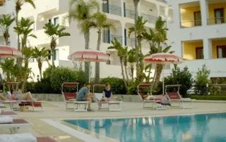 Außenaufnahme eines Hotelpools umringt von Palmen und Liegestühlen