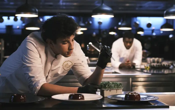 Yazid beim Anrichten eines Tellers, der junge Mann lehnt über drei Tellern, auf denen ein kleiner, runder Schokokuchen liegt, er hält eine Pinzette in der Hand. Im Hintergrund verschwommen wird er von einem weiteren Koch beobachtet.