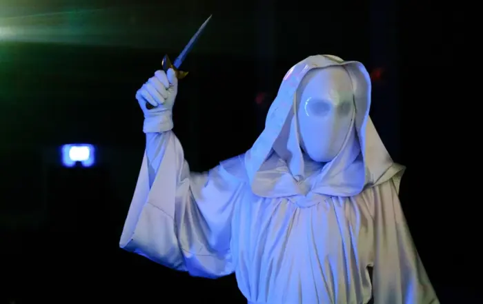 eine in eine weiße Kutte gekleidete Gestalt steht in einem dunklen Raum, sie trägt eine weiße Kapuze über ihrem maskierten Gesicht, sie hat die rechte Hand erhoben, in der sie einen Dolch hält