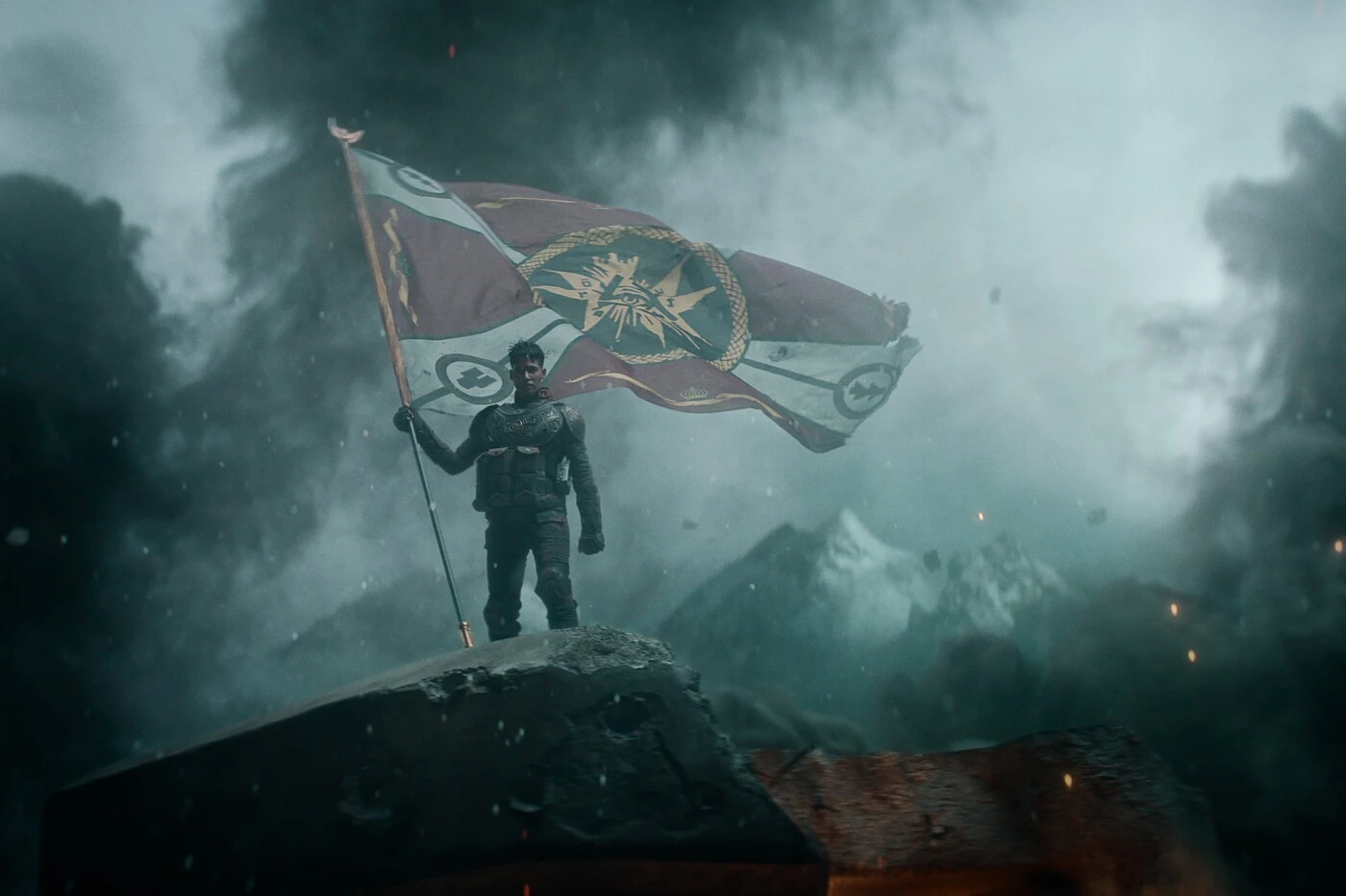 ein junger Mann in Rüstung steht auf der Spitze eines Felsens, er hält eine riesige Fahne in der Hand, auf der verschiedene Symbole abgebildet sind, im Hintergrund sind schneebedeckte Berge zu sehen, dunkler Rauch steigt um den Mann herum auf