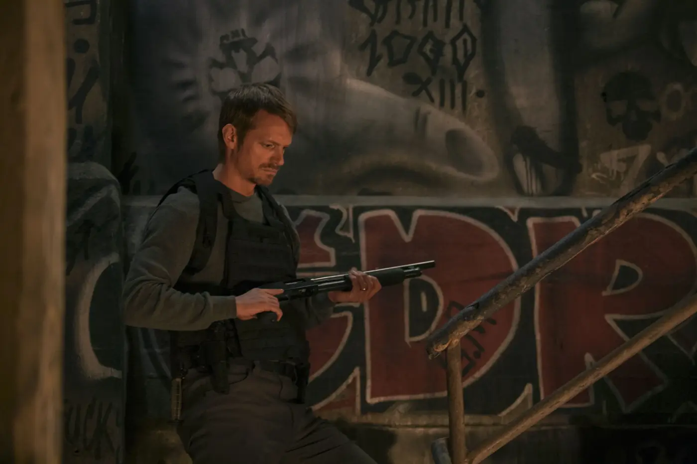 vor einer Wand, die mit Graffiti besprüht ist, steht ein Mann mit Schutzweste und einer Waffe in der Hand