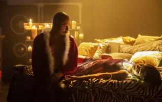 eine Frau, gekleidet in einem lilafarbenen Kleid und einer Lichterkette um den Hals, liegt auf einem Bett mit einem dutzend goldenen Kissen. Sie schaut zu einem Mann, der neben ihr sitzt und über seinem nackten Oberkörper eine weihnachtliche Jacke trägt. Sein kahler Kopf ist zerkratzt, während er zu der Frau sieht.