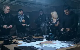 fünf Menschen in schwarzer Kleidung und schusssicheren Westen stehen in einem Lagerraum um einen Tisch, auf dem Tisch liegen Waffen, Munition und verschiedene Papiere