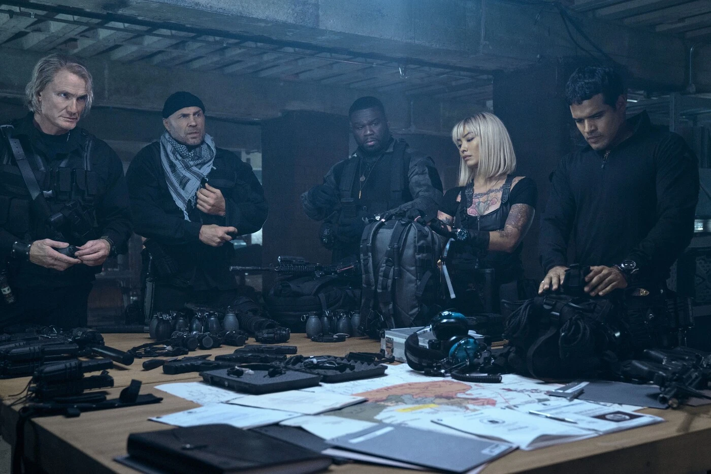 fünf Menschen in schwarzer Kleidung und schusssicheren Westen stehen in einem Lagerraum um einen Tisch, auf dem Tisch liegen Waffen, Munition und verschiedene Papiere