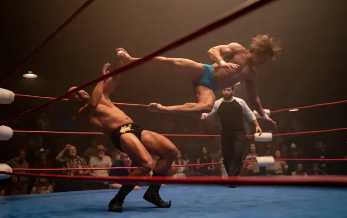 Wrestlingmatch, ein Mann in blauer Hose springt und führt einen Tritt aus, sein Gegner in schwarzer Hose stürzt zu Boden