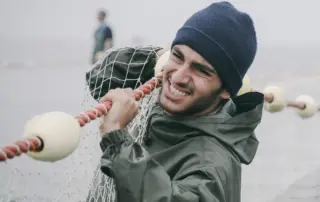 ein windiger Tag an der Küste, ein junger Mann in graugrünen Anorak und schwarzer Mütze zieht ein großes Netz aus dem Wasser