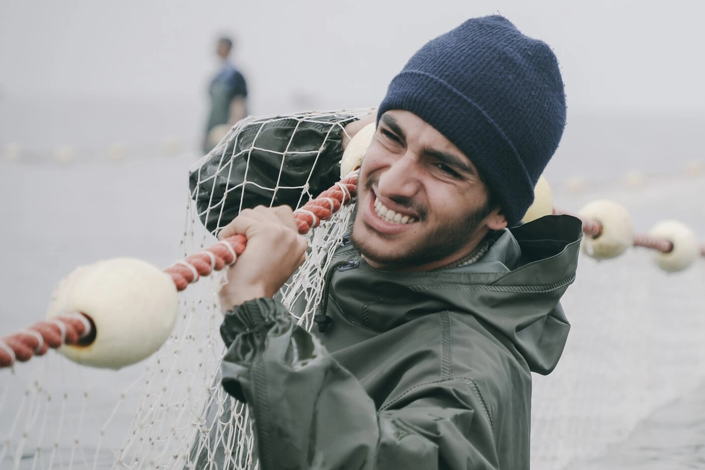 ein windiger Tag an der Küste, ein junger Mann in graugrünen Anorak und schwarzer Mütze zieht ein großes Netz aus dem Wasser
