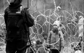 hinter einem Stacheldrahtzaun steht ein Mädchen in dicker Winterjacke und Mütze, sieht schaut zu einem bewaffneten Soldaten, der auf der anderen Seite des Zaunes mit dem Rücken zur Kamera steht