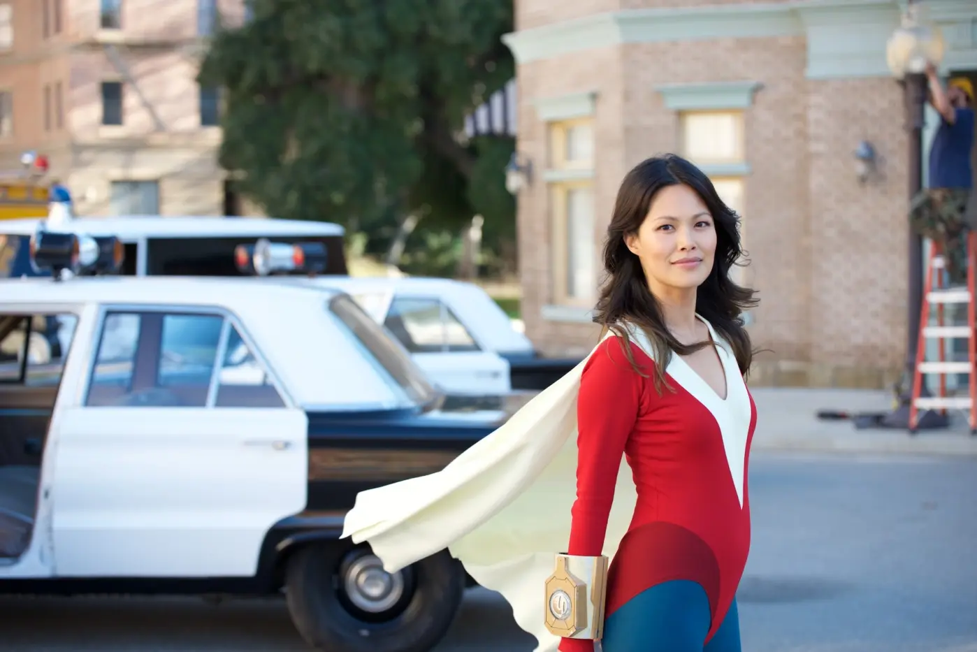 eine Straße bei Tag, im Hintergrund parken Polizeiautos, im Vordergrund steht eine junge Frau im altmodischen Superheldenanzug mit roten Oberteil, weißgelben Umhang und blauer Hose, sie lächelt in die Kamera