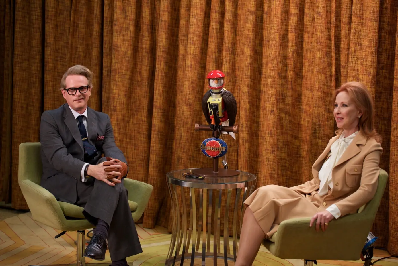 ein Fernsehinterview, vor einem braunen, altmodischen Vorhang sitzen ein Mann im grauen Anzug und eine Moderatorin, zwischen ihnen steht ein metallener Tisch auf dem ein Vogel mit einem roten Helm sitzt