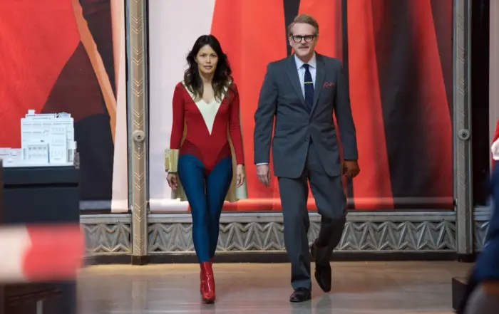 eine Frau im altmodischen Superheldenkostüm und ein Mann im Anzug gehen Seite an Seite durch einen Raum, im Hintergrund sind riesige Leinwände mit Gemälden zu erkennen, links ragt das Modell einer Stadt auf einem Tisch ins Bild