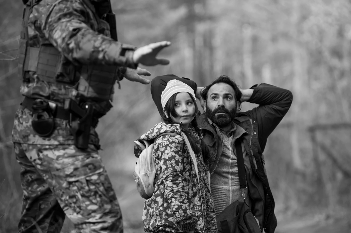im Hintergrund kniet ein Mann, die Hände hinter dem Kopf haltend, bei ihm steht seine Tochter. Ein Soldat, dessen Erscheinung im Vordergrund von den Bildrändern abgeschnitten wird, gibt ihnen Anweisungen