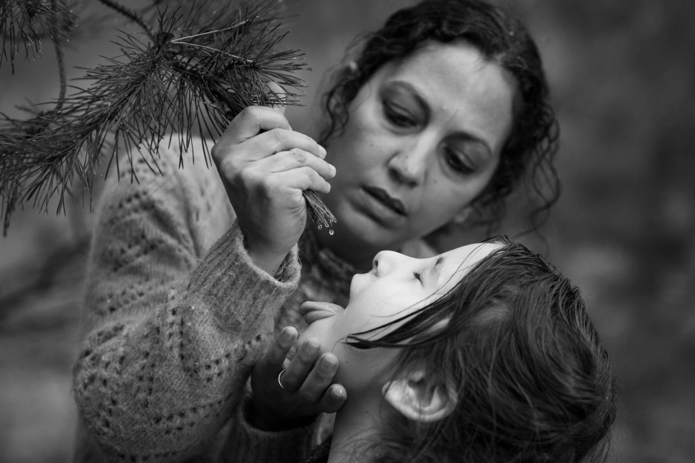 eine Frau hält einen Tannenzweig in ihrer rechten Hand von dem sie Wassertropfen in den Mund eines Mädchens tropft