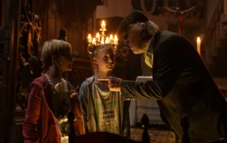 eine alte Villa bei Nacht, Kerzen erhellen Schränke und eine Treppe im Hintergrund, im Vordergrund stehen ein Junge, der Knoblauch um seinen Hals trägt, ein Mädchen, sowie ein älterer Herr, der mit seinem Zeigefinger belehrend auf den Jungen zeigt