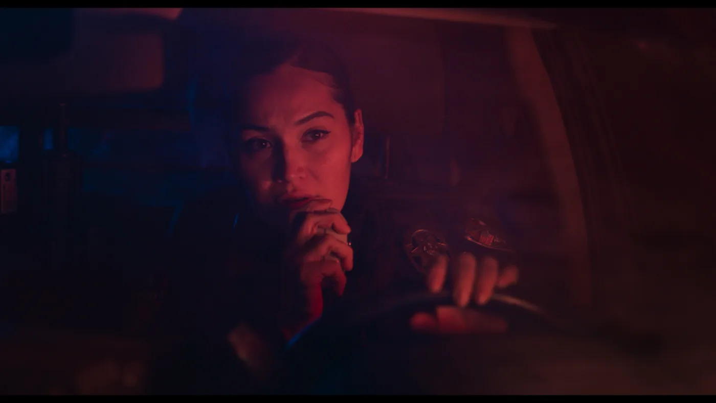 eine Polizistin sitzt in ihrem Auto hinter dem Lenkrad und spricht in ein Funkgerät, es ist Nacht, ihr Gesicht wird nur von einem roten Licht angeleuchtet