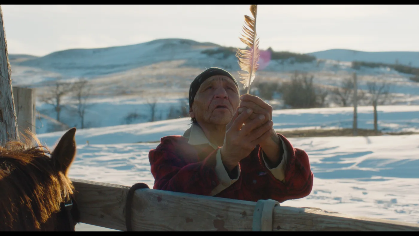 ein Mann steht am Zaun eines verschneiten Gatters, er hält eine Feder mit beiden Händen nach oben und betet, im Hintergrund ist eine verschneite Landschaft zu sehen