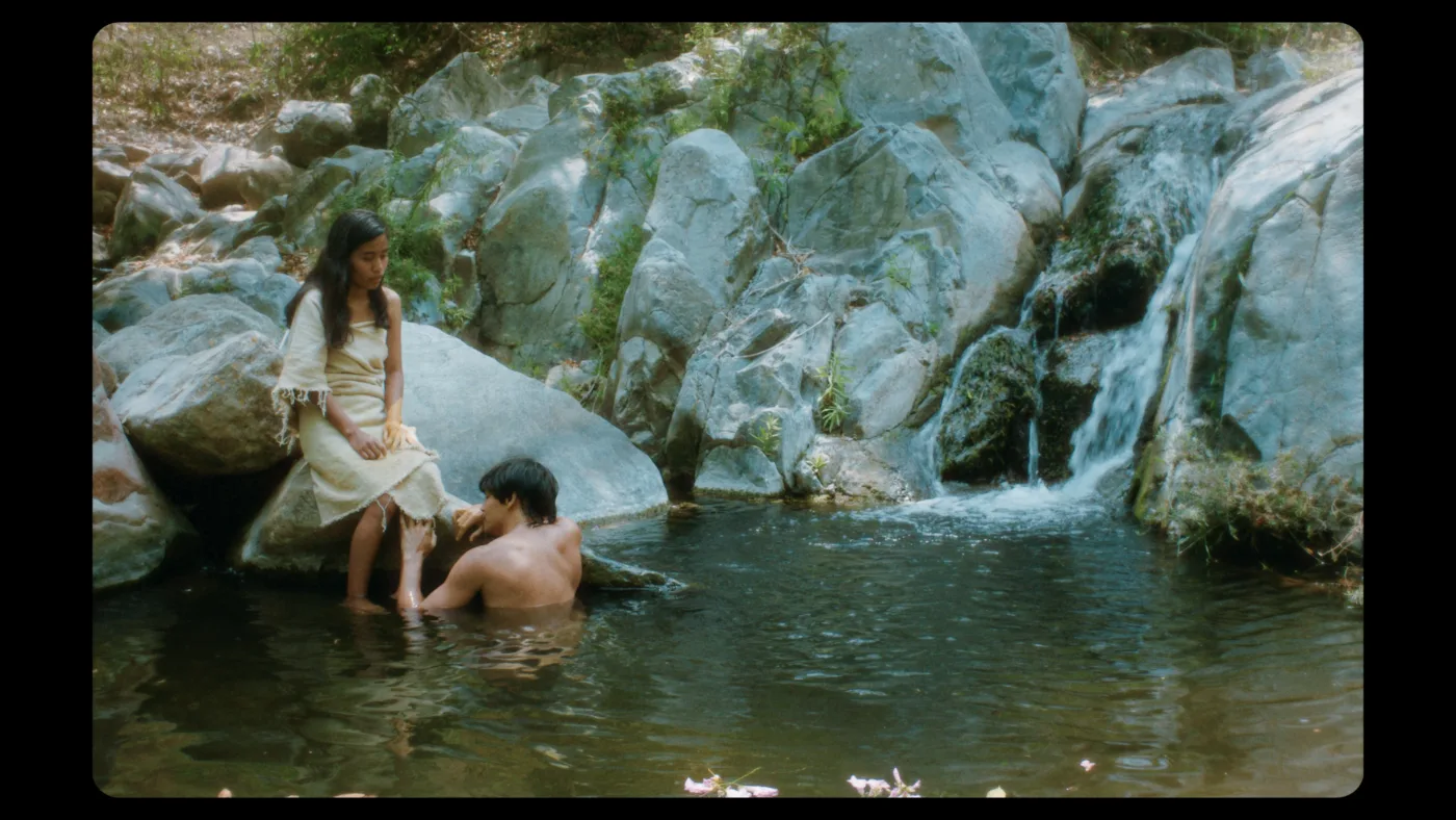 ein von kleinen Felsen umringter See im Urwald, links im Bild sitzt eine junge Frau auf einem großen Stein, vor ihm im Wasser ein oberkörperfreier Mann
