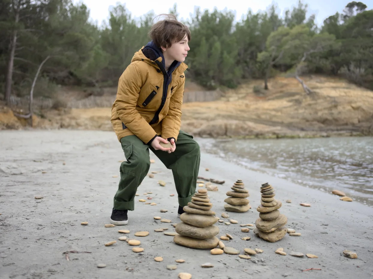 ein Junge steht am Ufer eines Gewässers, um ihn herum sind Türme aus Steinen gebaut, in den Händen hält er einen Stein