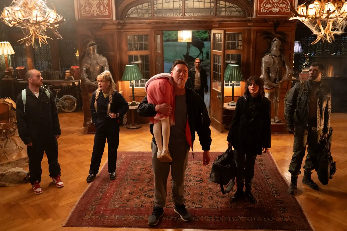 Vorsaal einer altmodisch eingerichteten Villa, sechs dunkel gekleidete Menschen betreten den Raum, mittig steht ein Mann, der ein Mädchen in weißer Strumpfhose und rosafarbenen Rock über seine Schulter trägt