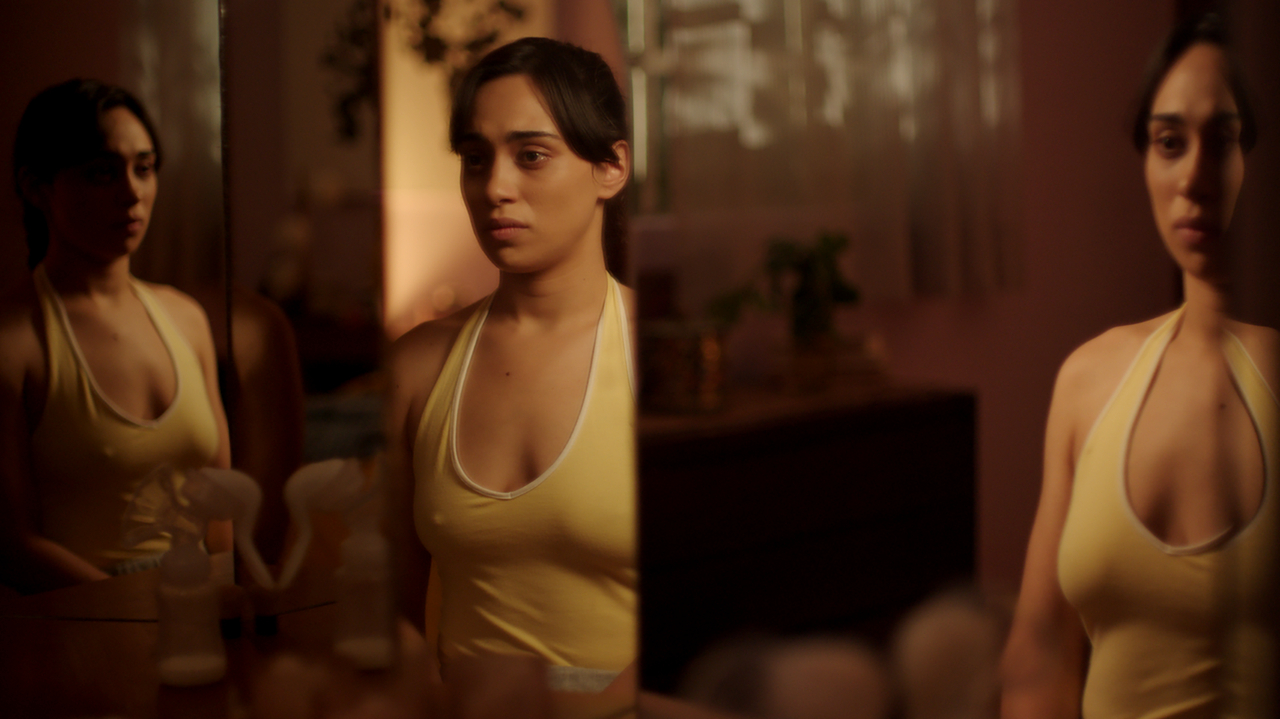 eine Frau im gelben Top steht vor einem mehrteiligen Wandspiegel in einer schummrigen Wohnung, ihr Spiegelbild doppelt sich