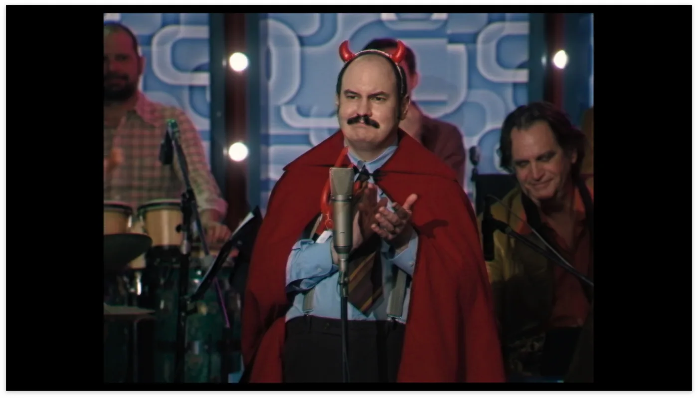 eine band tritt in einer Fernseh-Kulisse auf, im Hintergrund sind Musiker an verschiedenen Instrumenten zu sehen, in der Mitte steht ein Mann im Hemd und Krawatte am Mikrofon, er trägt einen roten Umhang und einen Haarreif mit kleinen Teufelshörnern