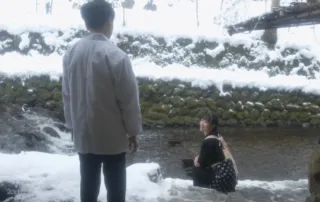 eine Frau in Hoteluniform sitzt am Ufer eines schmalen Flusses, sie dreht sich zu einem Mann, der mit dem Rücken zur Kamera steht und mit ihre redet, es schneit