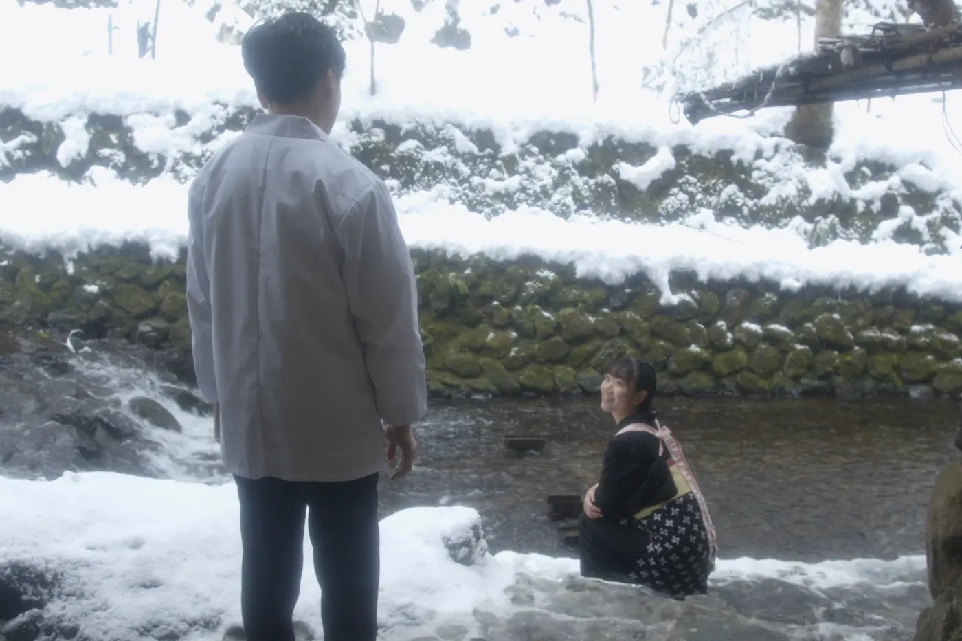eine Frau in Hoteluniform sitzt am Ufer eines schmalen Flusses, sie dreht sich zu einem Mann, der mit dem Rücken zur Kamera steht und mit ihre redet, es schneit