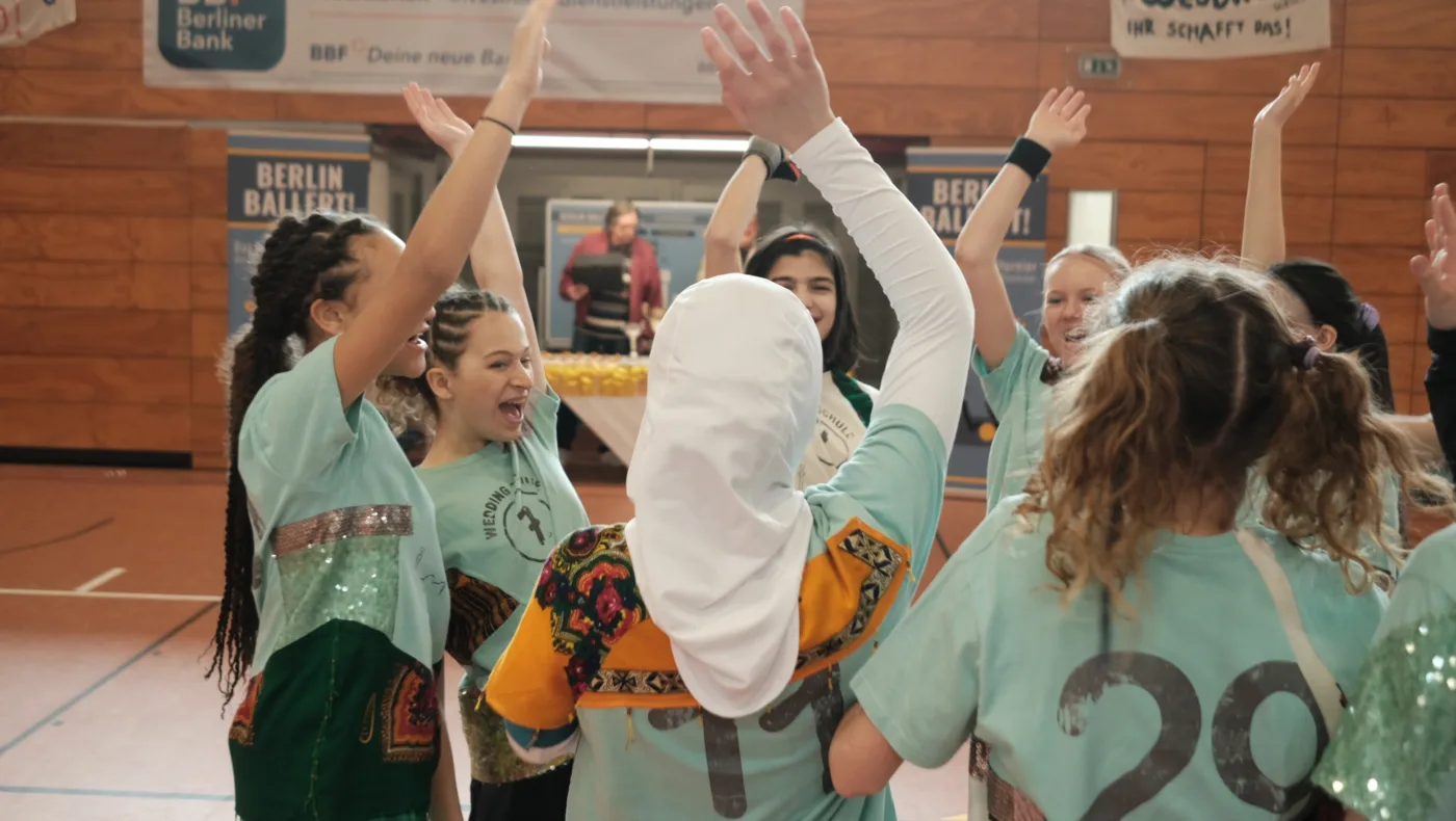 im Inneren einer Sporthalle, ein Team aus jugendlichen Mädchen, gekleidet in Sportsachen und hellblaue Leibchen steht im Kreis und reißt die Arme nach oben