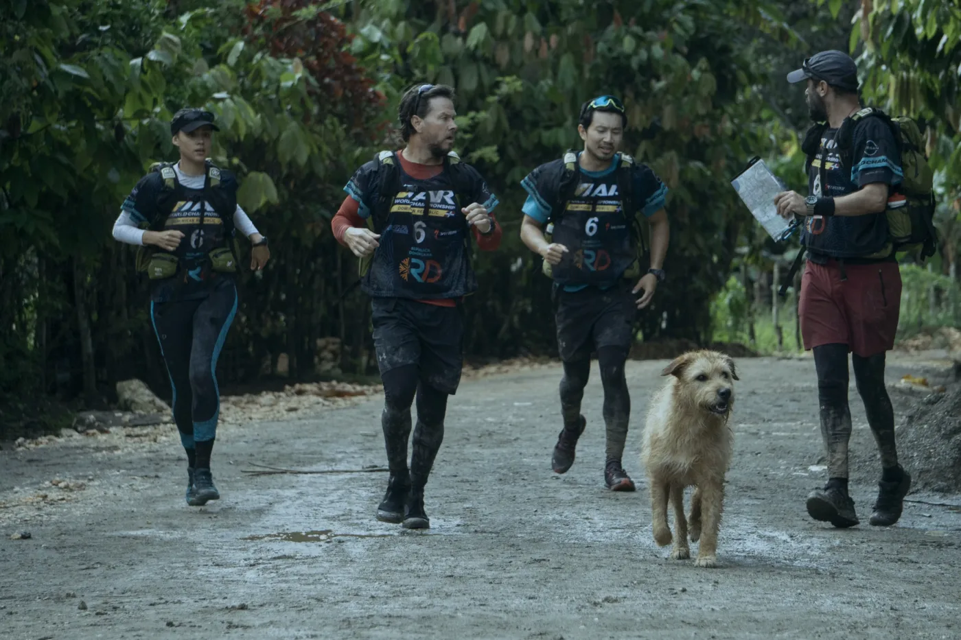 vier Athleten rennen auf einem Weg durch eine bewaldete Gegend, zwischen ihnen läuft ein Hund