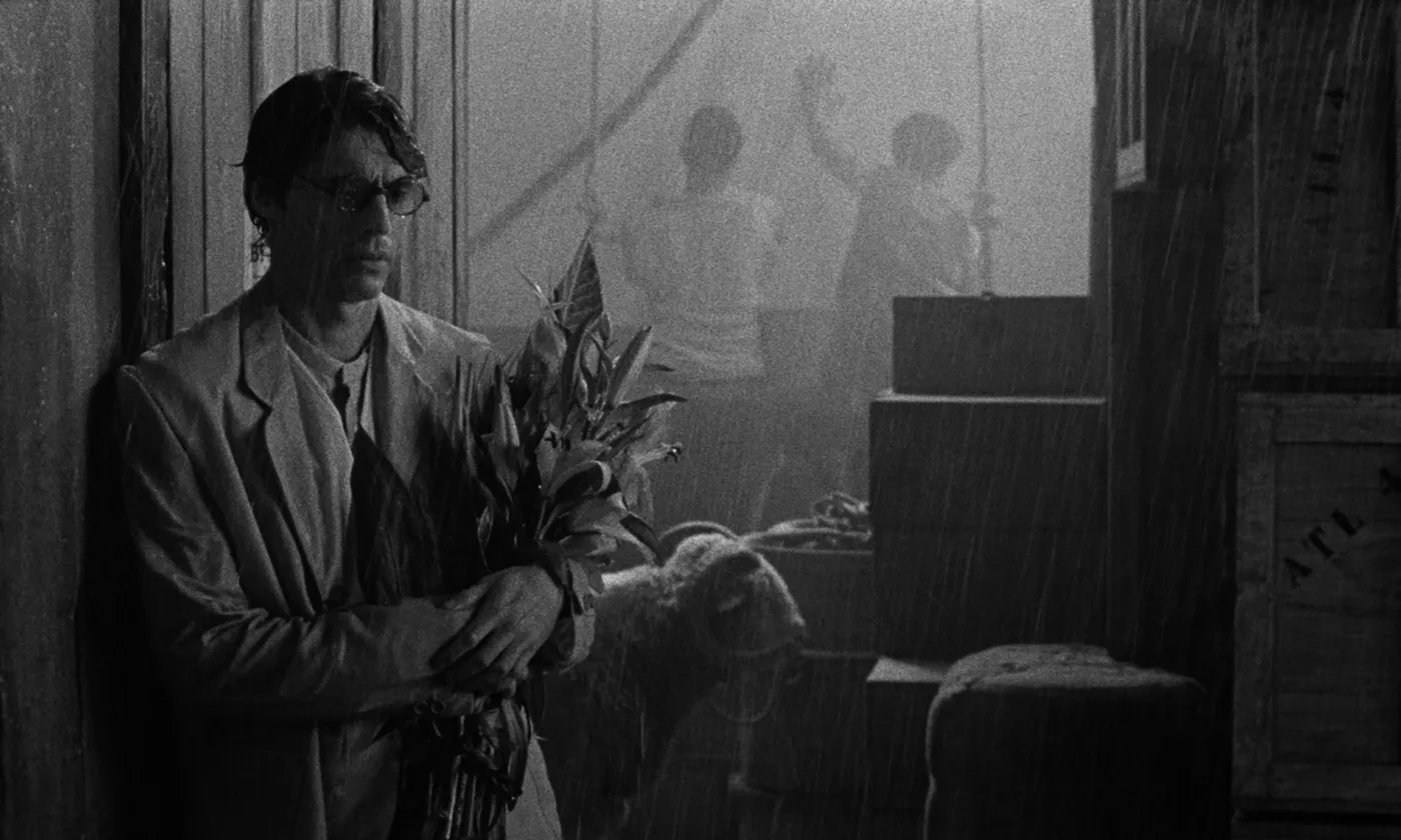 schwarzweiß Aufnahme eines Mannes, der mit einem Blumenstrauß im Arm im Regen vor einer hölzernen Wand steht, neben ihm stapeln sich Kisten in die Höhe, im Hintergrund arbeiten zwei weitere Männer an einer Wand