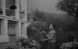 schwarzweiß Aufnahme eines kleinen Gartens, in dem eine junge Frau die Pflanzen gießt, während sie zu einem Balkon aus weißem Stein am rechten Bildrand aufsieht, von dem eine weitere Frau blickt