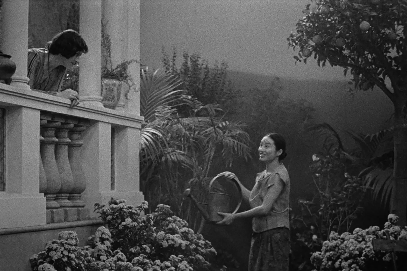 schwarzweiß Aufnahme eines kleinen Gartens, in dem eine junge Frau die Pflanzen gießt, während sie zu einem Balkon aus weißem Stein am rechten Bildrand aufsieht, von dem eine weitere Frau blickt