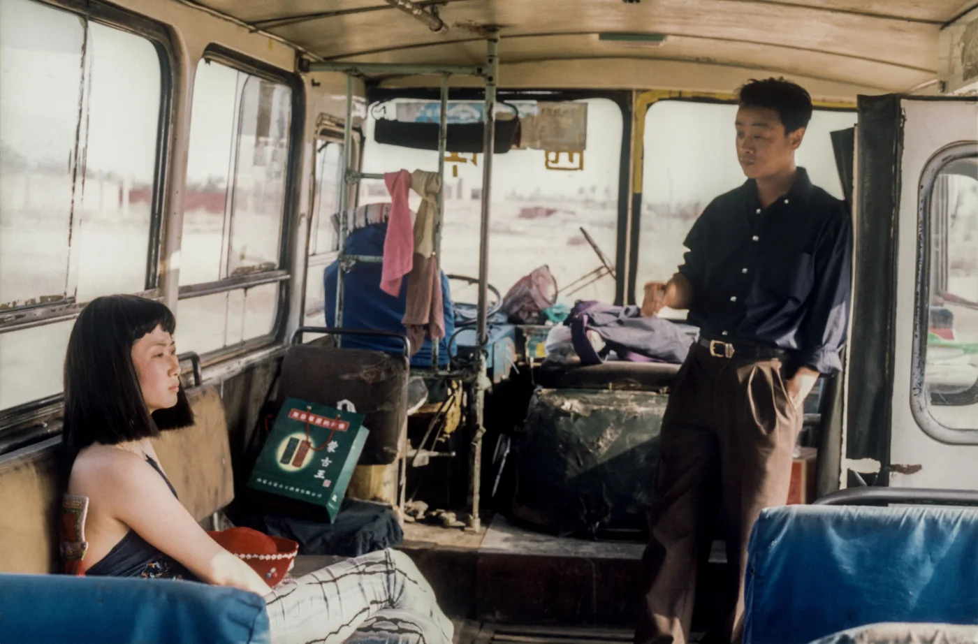 Innenaufnahme eines ausrangierten Busses, links sitzt eine Frau auf einer Bank, rechts steht ein Mann an der Tür, im Hintergrund verteilt sich Kleidung