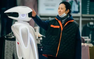 eine Frau in dunkler Jacke mit hellorangen Reißverschluss legt ihre Hand auf einen links im Bild stehenden, weißen, futuristischen Roboter