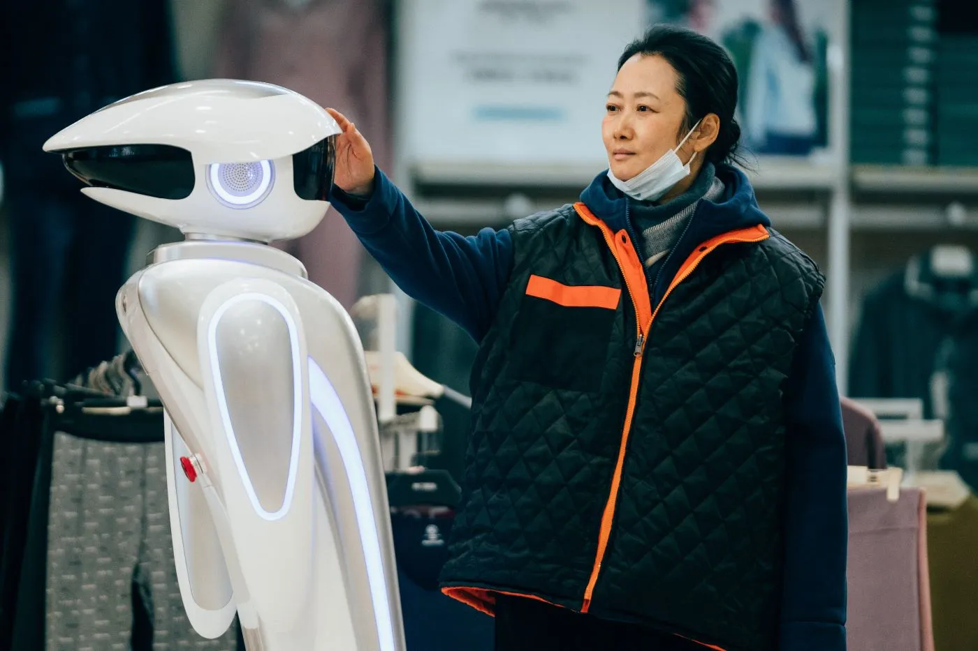 eine Frau in dunkler Jacke mit hellorangen Reißverschluss legt ihre Hand auf einen links im Bild stehenden, weißen, futuristischen Roboter