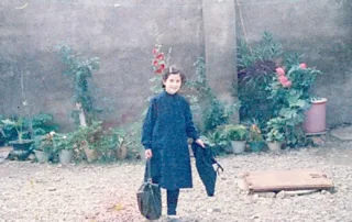 ein Foto der Regisseurin in Kinderjahren, sie steht vor einer grauen Steinmauer und einigen Pflanzen, sie lächelt in die Kamera während sie eine Tasche in der rechten Hand trägt