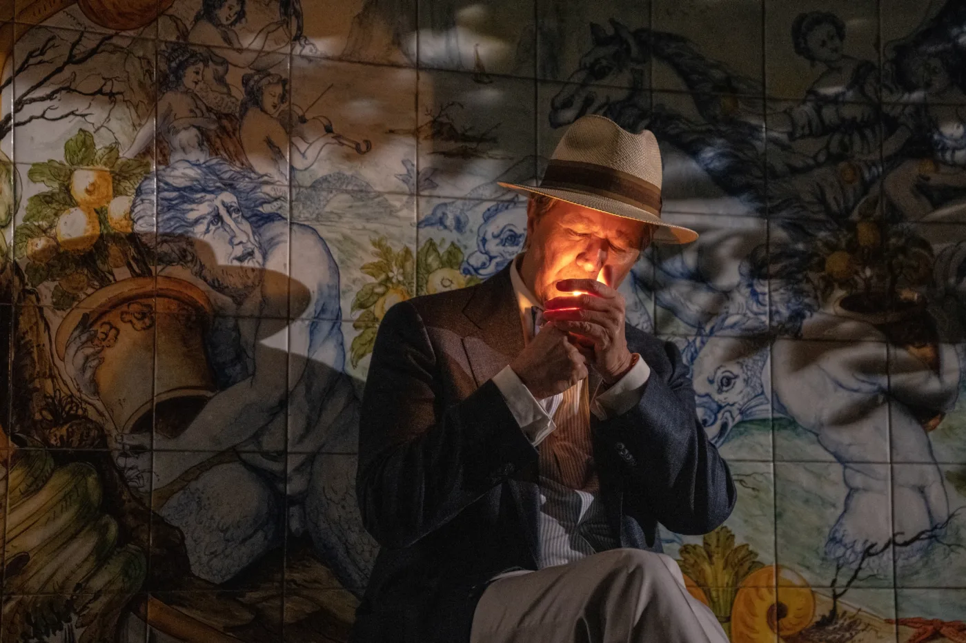 halbnahe Aufnahme eines Mannes mit Anzug und Hut, der sich vor einem Gemälde auf Wandfliesen eine Zigarette anzündet