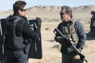 zwei bewaffnete Männer in schusssicheren Westen und mit Sonnenbrille unterhalten sich vor karger Landschaft