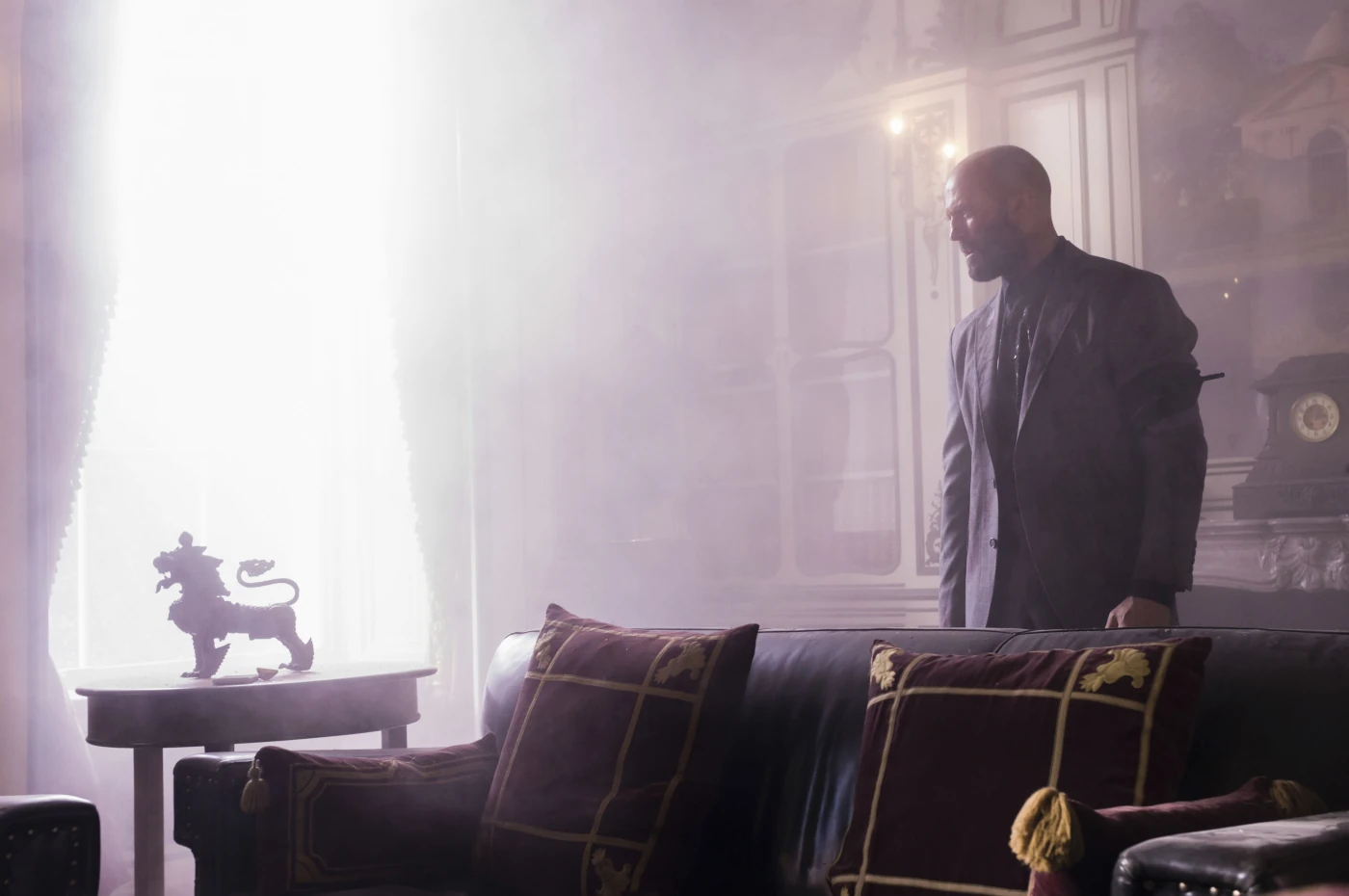 ein elegant ausgestattetes Zimmer, Licht aus einem großen Fenster blendet die Kamera, im rechten Teil des Bildes steht Jason Statham im Anzug hinter einem schwarzen Sofa