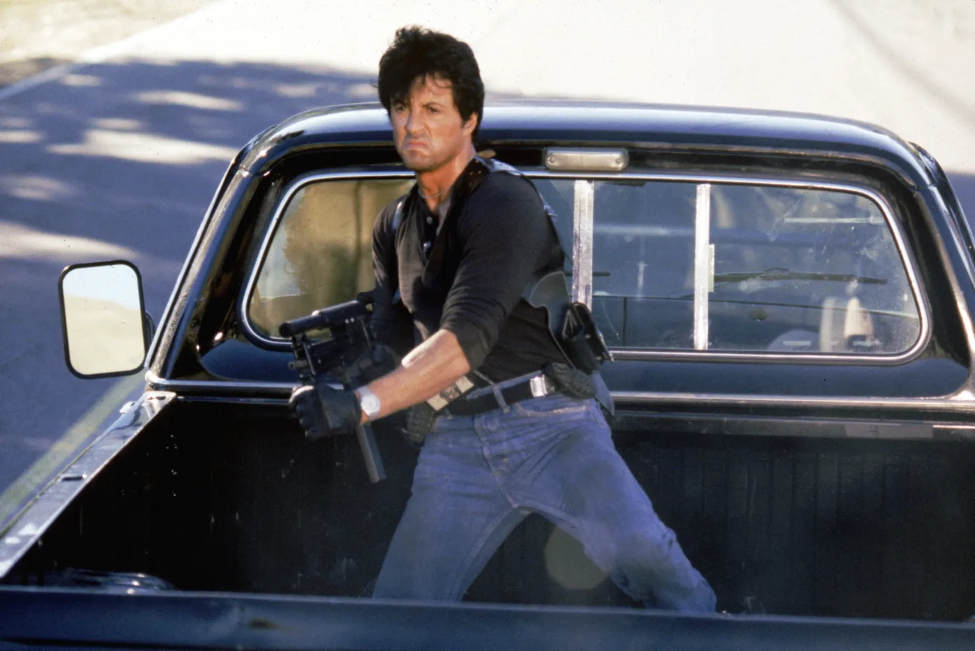 ein schwarzer Wagen mit Ladefläche fährt auf einer Straße von der Kamera weg, auf der Ladefläche steht Sylvester Stallone mit einem Maschinengewehr bewaffnet und schießt auf Verfolger außerhalb des Bildes