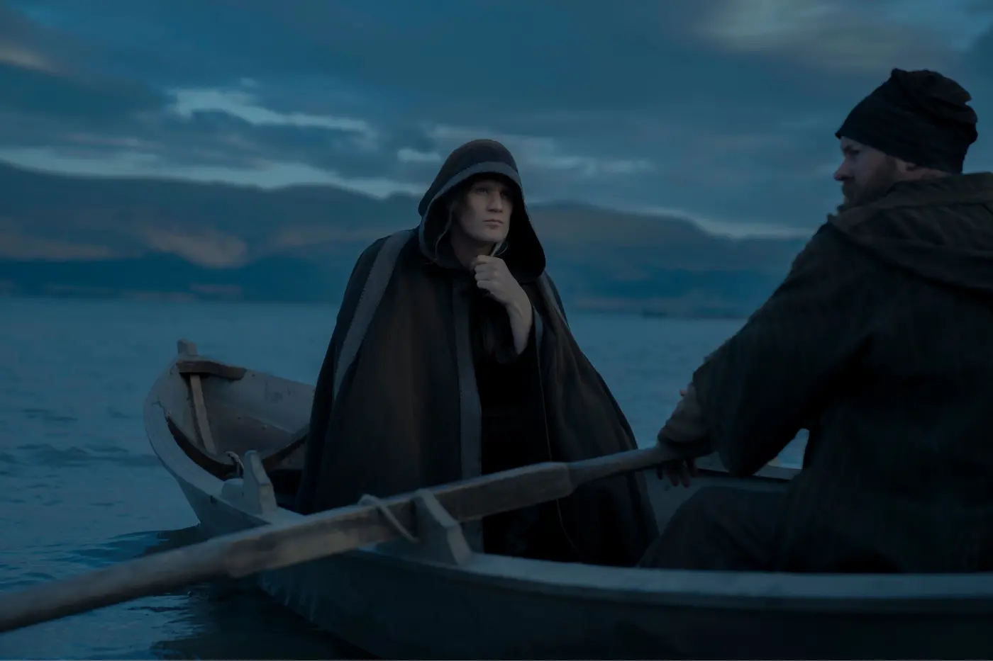 in der Dämmerung fährt ein kleines Boot über Wasser, im Hintergrund ist Land zu sehen, im Boot sitzt ein Ruderer und ein in einen schwarzen Umhang gekleideter Daemon Targaryen