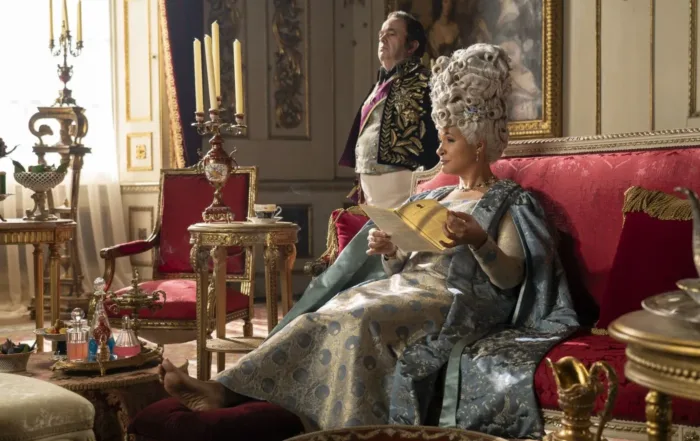 Innenaufnahme eines edel möblierten Zimmers, eine Frau mit hochgesteckten weißen Haaren sitzt auf einem Sofa und blättert in einer kleinen Zeitung, hinter ihr steht ein Bediensteter