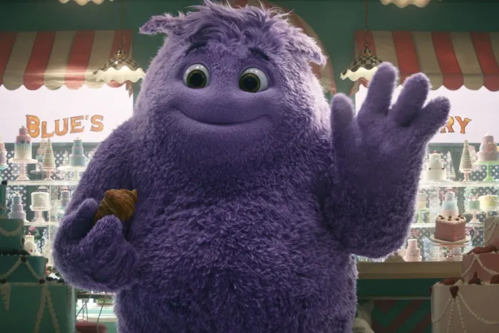 im Inneren einer Bäckerei, links und rechts stehen verzierte Torten, die Mitte des Bildes füllt ein großes Wesen mit lilafarbenen Fell, welches mit der einen Hand winkt und in der anderen Hand ein Stück Kuchen hält